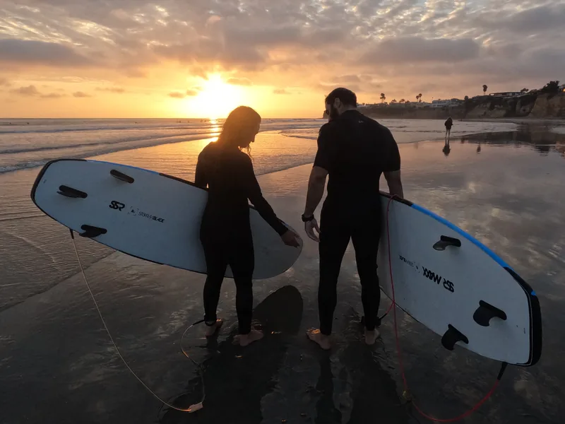 Two surfers carrying their boards along the shoreline at sunset